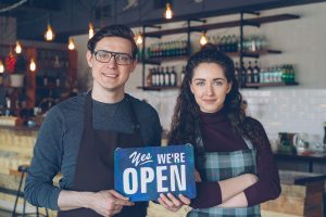 Best Small Business Marketing Services Jensen Beach Florida a Man and a Woman Standing Inside a Cafe Holding a Sign That Says Yes, We're Open